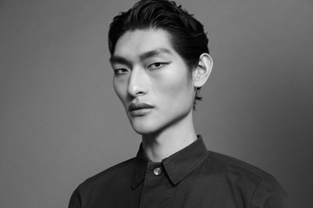 Monochrome portrait of an adult Asian man with slicked-back hair and a button-down shirt in a studio environment with diffused lighting.