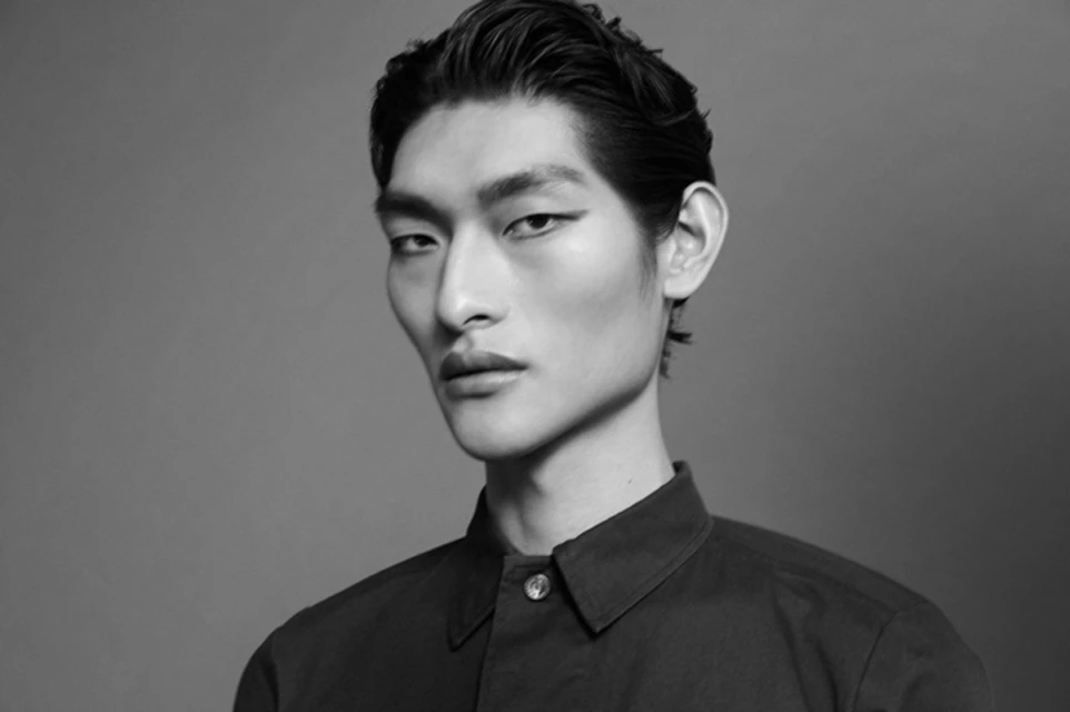 Monochrome portrait of an adult Asian man with slicked-back hair and a button-down shirt in a studio environment with diffused lighting.