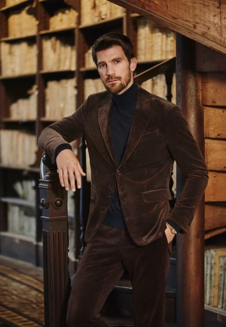 Man in brown velvet suit poses near wooden staircase and bookshelf, showcasing classic style in an indoor setting with warm lighting.