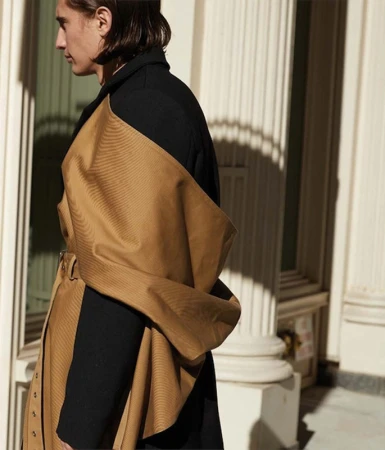 Profile of a man wearing a black coat draped with a camel-colored fabric panel against classical columns in Los Angeles.