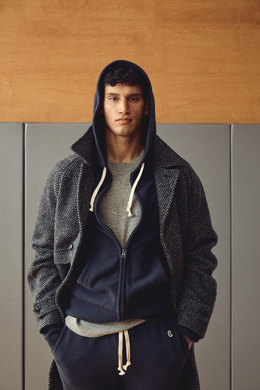Portrait of a young adult male with curly dark hair wearing a grey t-shirt, blue hooded sweatshirt, herringbone overcoat, and blue sweatpants.