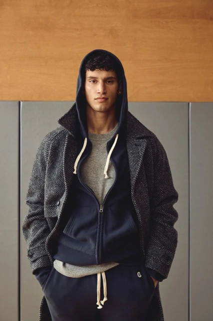 Portrait of a young adult male with curly dark hair wearing a grey t-shirt, blue hooded sweatshirt, herringbone overcoat, and blue sweatpants.