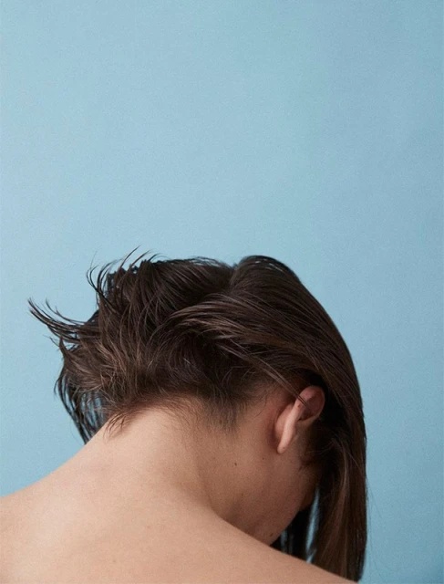 From behind, a woman with dark hair and an undercut poses against a light blue background in a close-up studio portrait.