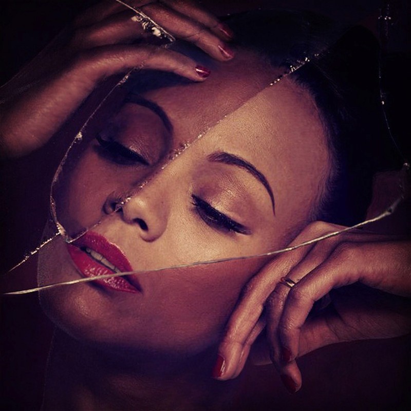 Portrait of an adult female with hands near her face and red lipstick, seen through broken glass fragments against a dark background.