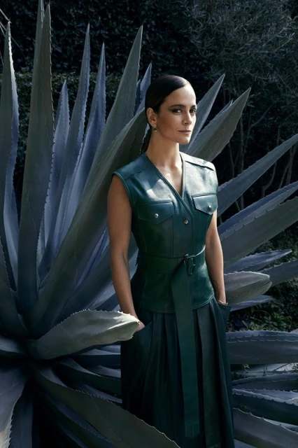 Adult woman in a belted green leather dress and sleeveless jacket posing in front of a tall agave plant in an outdoor setting.