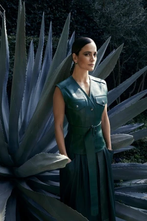 Adult woman in a belted green leather dress and sleeveless jacket posing in front of a tall agave plant in an outdoor setting.