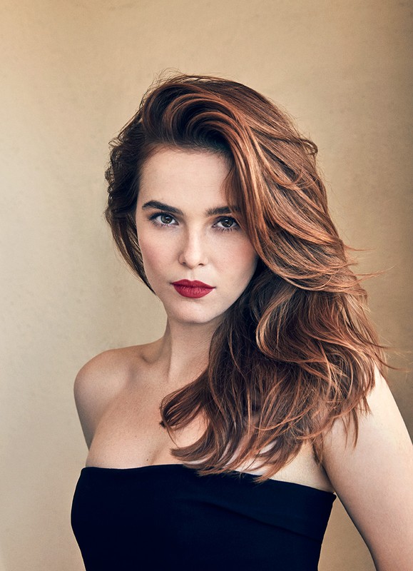 Portrait of a woman with long brown hair, dark lipstick, wearing a black strapless top against a neutral-toned background in soft lighting.