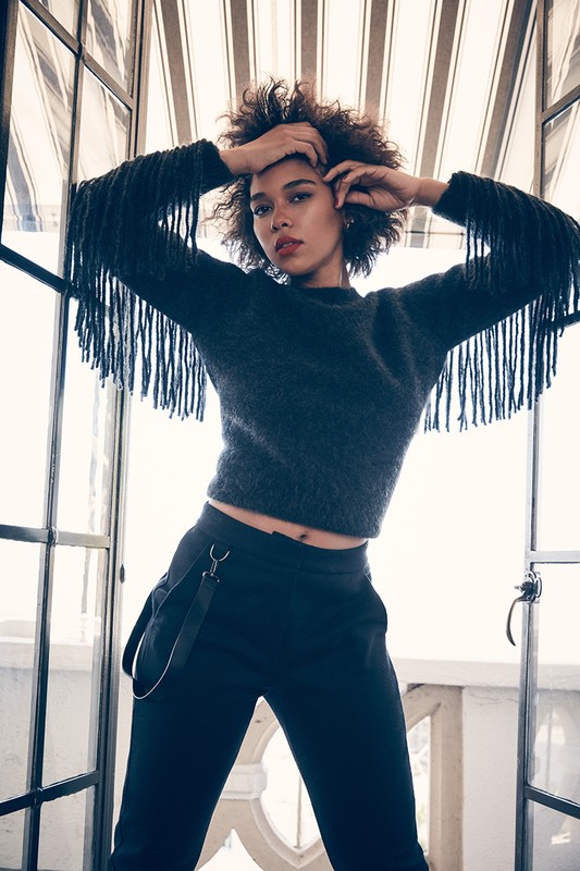 Fashionable woman with afro poses in a gray fringe sweater and black pants against a bright, framed window, showcasing style and confidence.