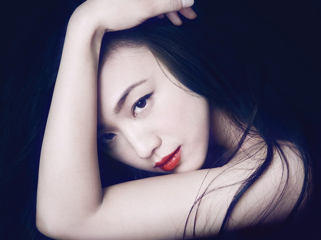 Portrait of an Asian woman with dark hair and bright red lipstick, resting her arms behind her head against a dark background.