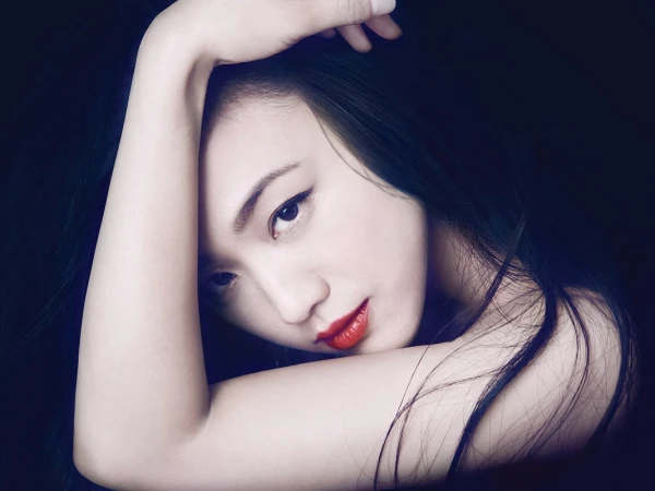 Close up portrait of a woman with dark hair and red lipstick resting her head on her raised arm against a dark background.