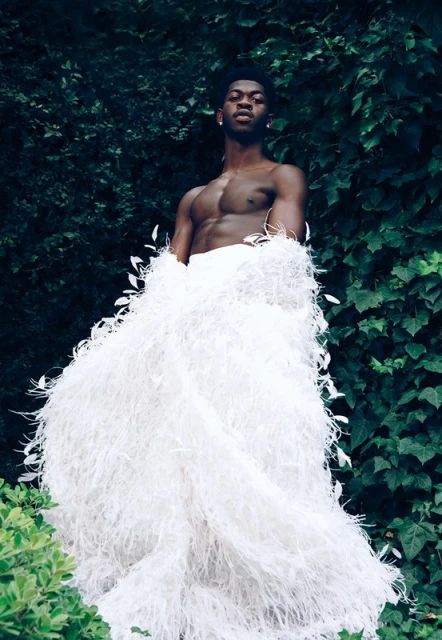 A bare-chested man in a white, feathery garment poses outdoors amid lush greenery, striking a confident pose in natural light.