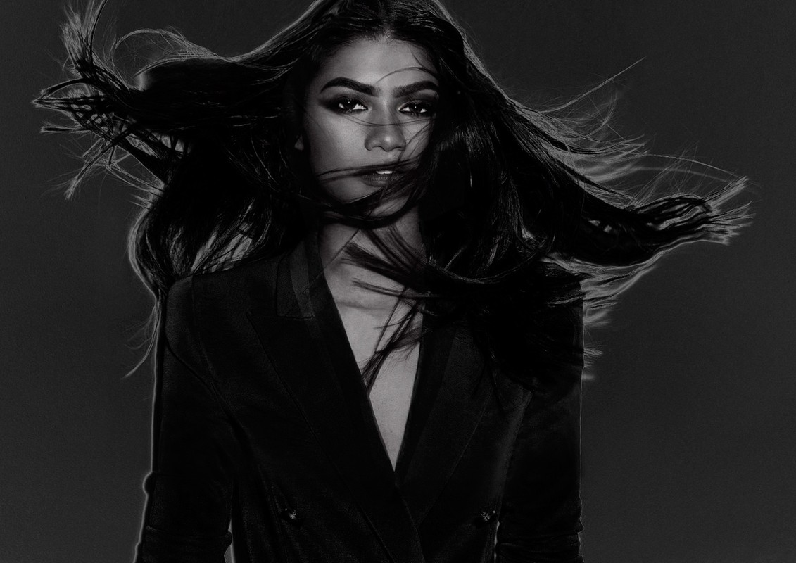 Portrait of a woman with long dark hair blowing across her face wearing a dark blazer, photographed in black and white by James White.