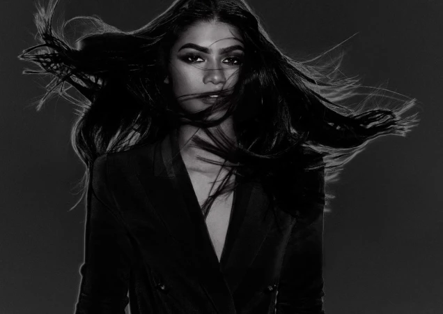Portrait of a woman with long dark hair blowing across her face wearing a dark blazer, photographed in black and white by James White.