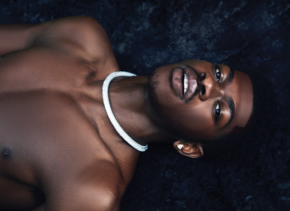 Close-up portrait of a shirtless Black man wearing a thick silver diamond choker necklace against a dark textured background.