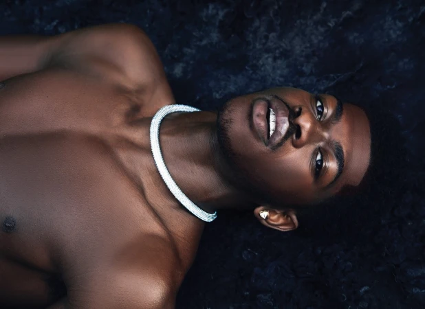 Close-up portrait of a shirtless Black man wearing a thick silver diamond choker necklace against a dark textured background.