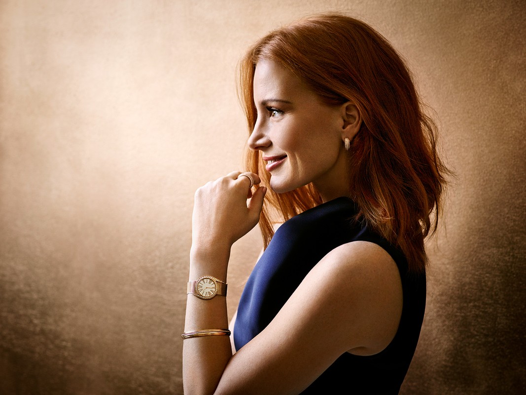 Elegant studio portrait of a redhead woman in a dark dress, wearing gold jewelry, smiling softly in profile against a textured backdrop.