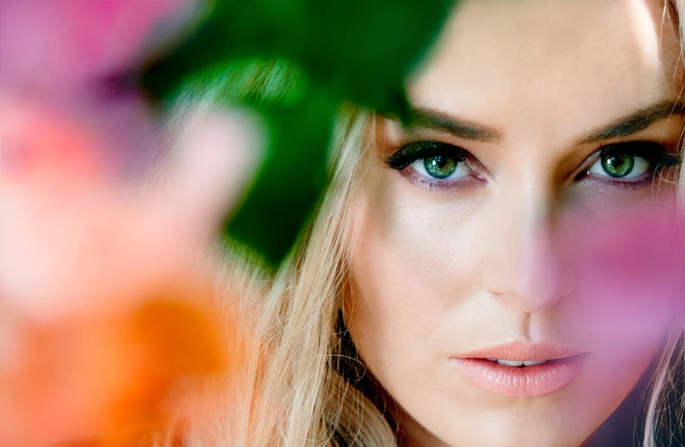 Close-up portrait of a blond adult female with green eyes, soft makeup, and pink floral elements in the foreground.