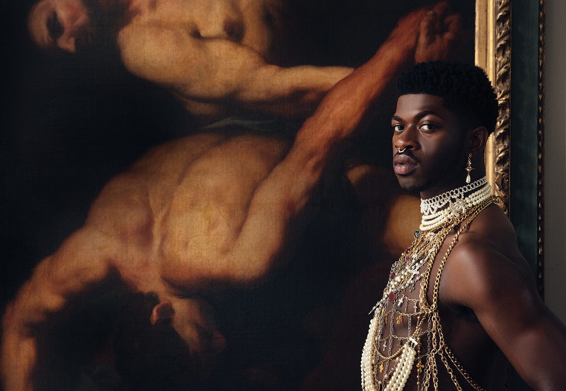 Lil Nas X poses indoors with a historical painting, wearing jewelry including a pearl choker and gold chains in a portrait shot.