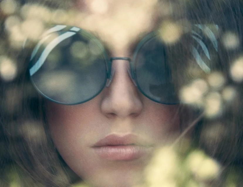 Close-up portrait of a woman wearing round sunglasses, soft focus, and blurred floral accents in the foreground and background.