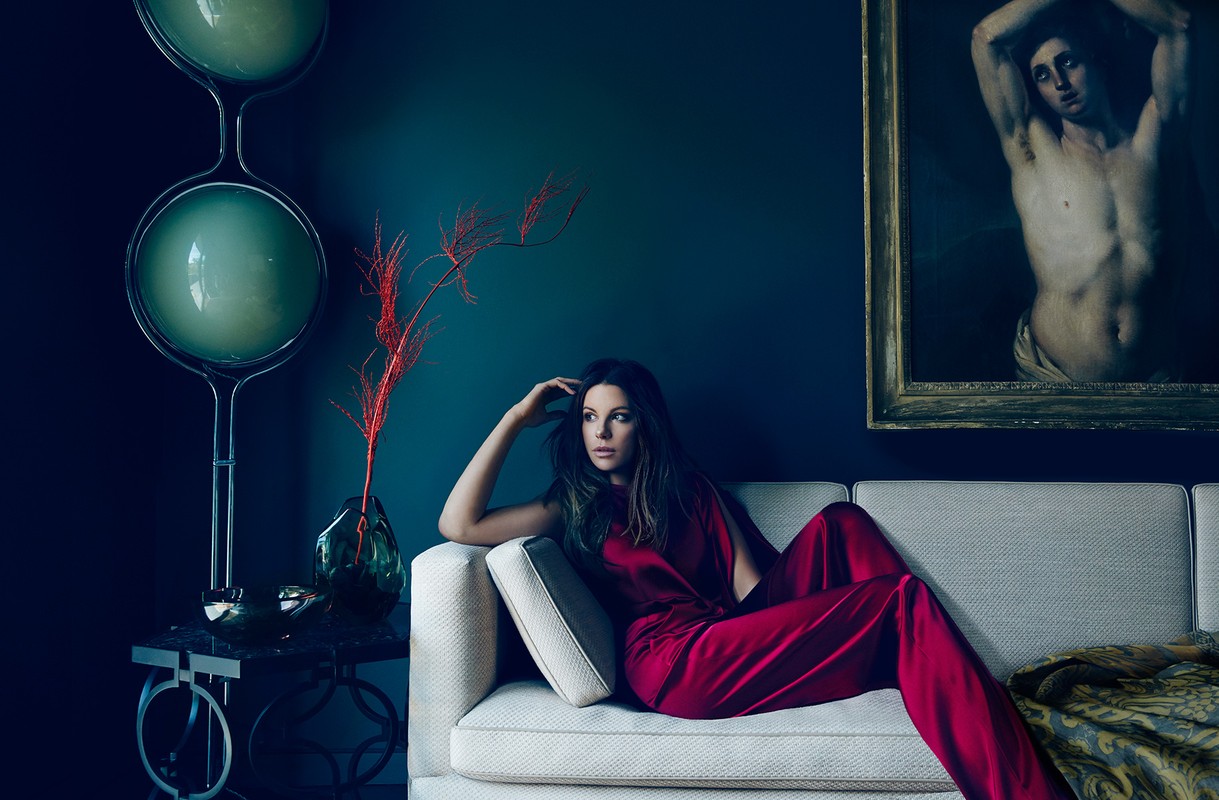 Adult woman in red jumpsuit lounges on cream couch in a home interior with art, a modern lamp, and teal walls in a lifestyle fashion shot.