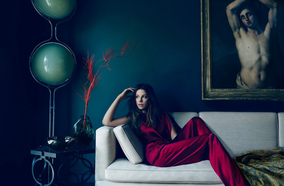 Adult woman in red jumpsuit lounges on cream couch in a home interior with art, a modern lamp, and teal walls in a lifestyle fashion shot.