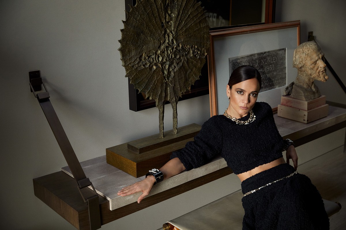 Woman with dark hair and stylish black outfit poses indoors, wearing jewelry, next to artwork and furniture in a staged environment.