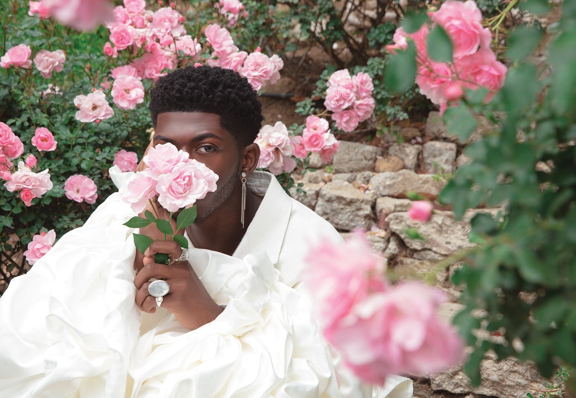 Portrait of an African American man holding pink roses, wearing a white ruffled top, with more pink roses and stone wall background.