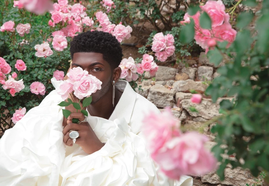 Portrait of an African American man holding pink roses, wearing a white ruffled top, with more pink roses and stone wall background.