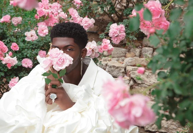 Portrait of an African American man holding pink roses, wearing a white ruffled top, with more pink roses and stone wall background.
