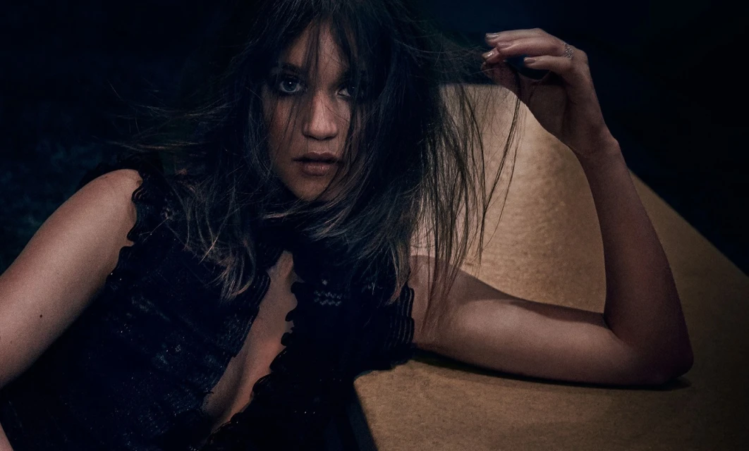 Close-up portrait of a woman with dark hair, wearing a black top, looking at the camera, with a neutral expression in a dimly lit indoor space.