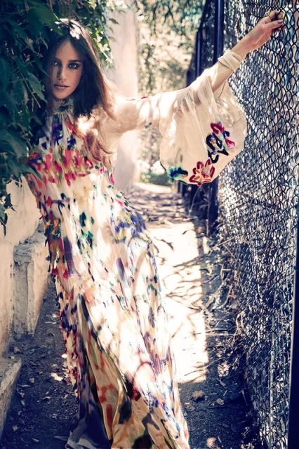 A woman stands by a metal fence wearing a long dress with a colorful floral print and bell sleeves, surrounded by green foliage.