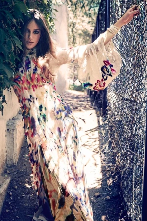 A woman stands by a metal fence wearing a long dress with a colorful floral print and bell sleeves, surrounded by green foliage.