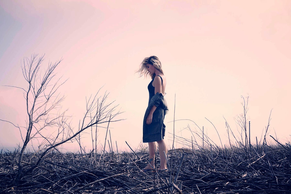 Adult woman standing barefoot in a black dress with a jacket over her shoulder in a field of dried twigs, pale pink sky.