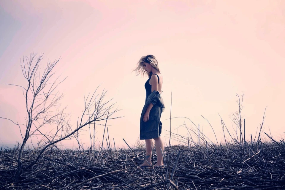 Adult woman standing barefoot in a black dress with a jacket over her shoulder in a field of dried twigs, pale pink sky.