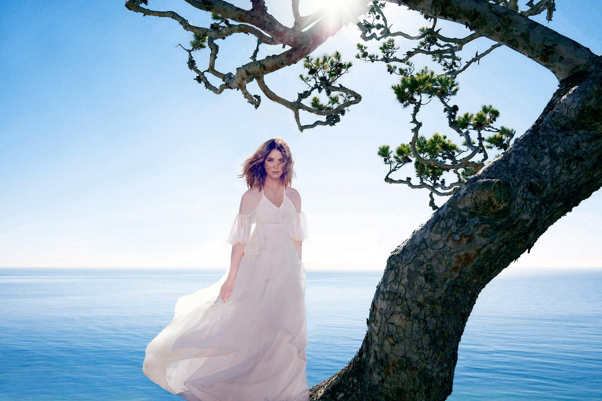 Woman in a flowing white dress standing outdoors near a tree branch with the ocean and clear sky in the background.