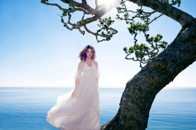 Woman in a flowing white dress standing outdoors near a tree branch with the ocean and clear sky in the background.