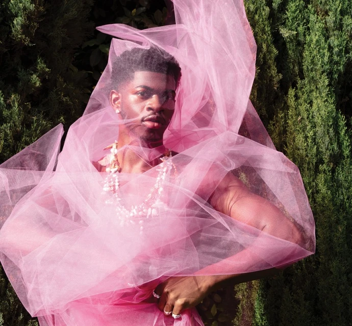 Portrait of an adult male posing outdoors surrounded by a pink tulle garment and wearing a pearl necklace against a greenery backdrop.