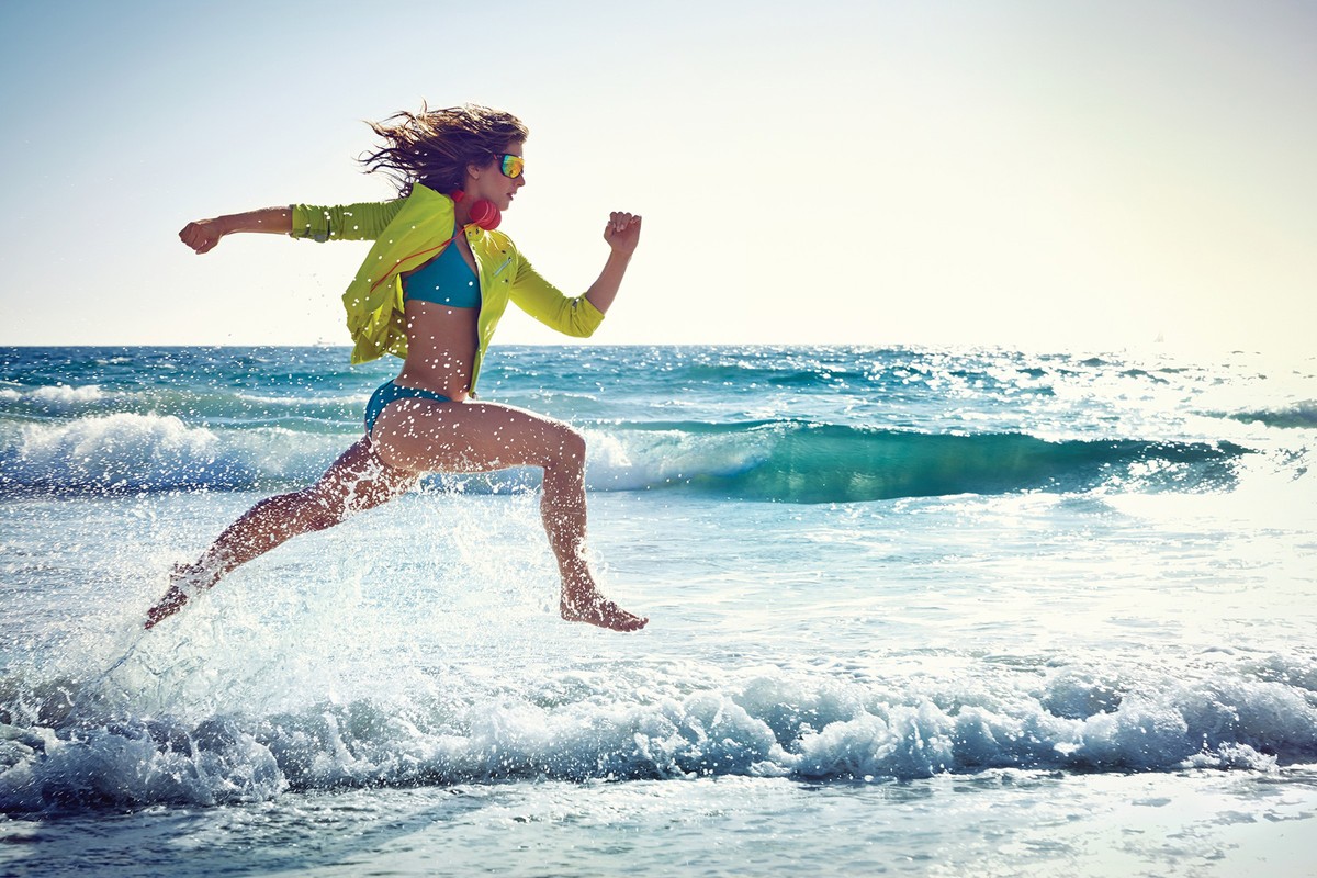 Woman in a yellow sports jacket and blue bikini running through ocean surf along a sandy beach