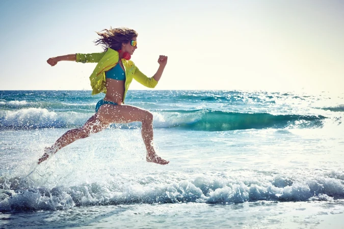 Woman in a yellow sports jacket and blue bikini running through ocean surf along a sandy beach