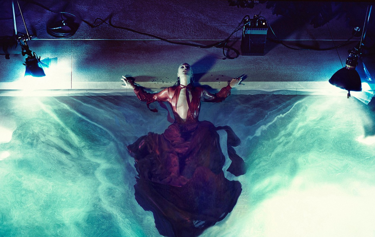 An adult woman lies in water wearing a red dress, with lights overhead. The scene is lit with blue and white lights.