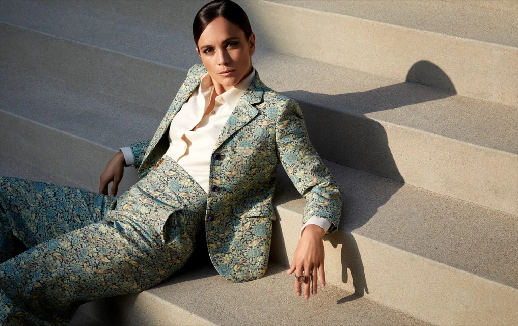 Woman in a floral suit sitting on concrete steps, looking at the camera. She wears a white button-up shirt with rings on her fingers.