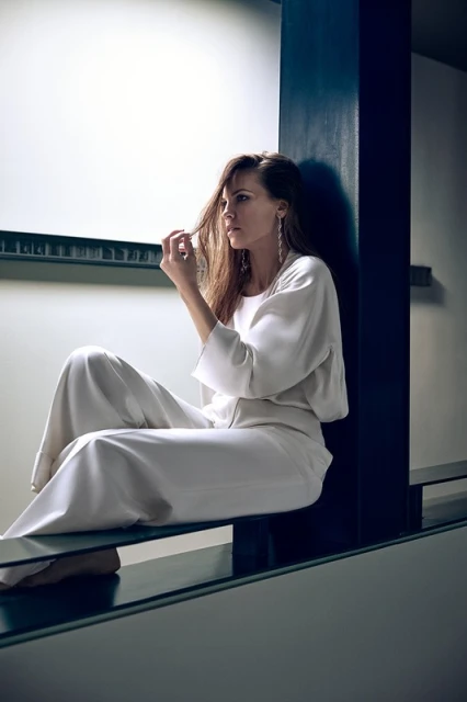 Woman in white pantsuit sits on a glass ledge near a dark column in an indoor setting, looking away with a pensive expression.
