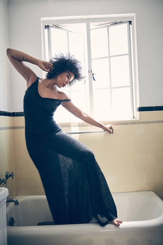 Woman in black dress poses in a bathroom with a window in the background. The scene is brightly lit and clean.