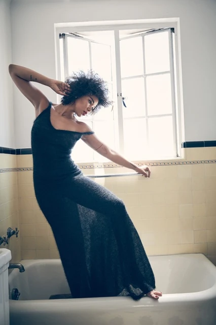 Woman in black dress poses in a bathroom with a window in the background. The scene is brightly lit and clean.