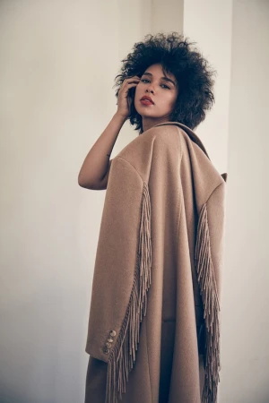 Woman with afro poses wearing a tan fringed coat looking over her shoulder in a fashion studio shot. Minimalist aesthetic.