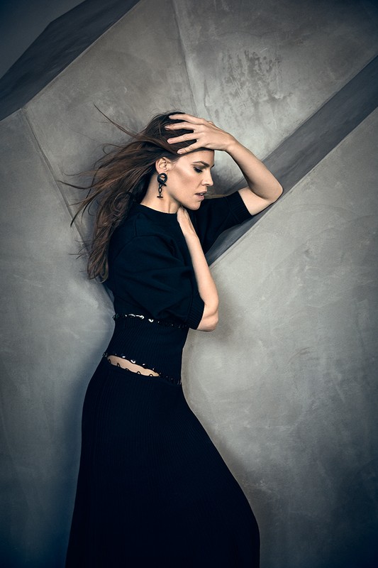 Woman in a black dress with long hair blowing, posing against a textured grey concrete wall.