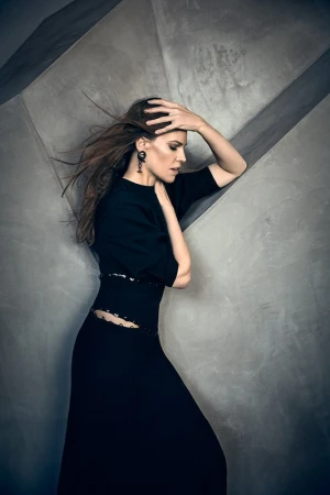 Woman in a black dress with long hair blowing, posing against a textured grey concrete wall.