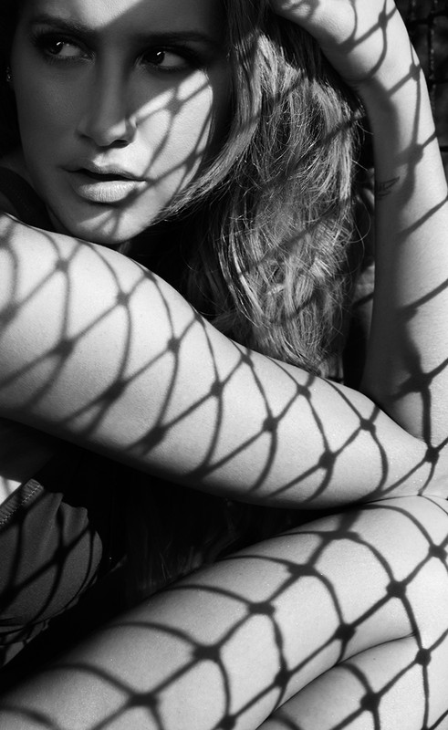 Black and white portrait of a woman partially covered by a net with shadows on her arms and face in an indoor setting.