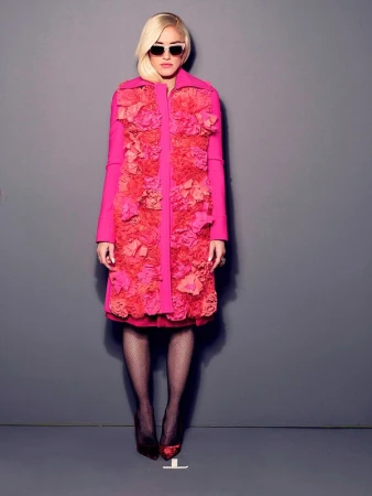 Woman in dark sunglasses wearing a vibrant pink textured floral coat and fishnet tights against a grey backdrop