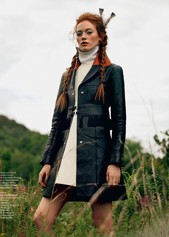 A model with red hair in pigtails poses outdoors, wearing a black leather coat, white turtleneck, and feathers in her hair, in a fashion editorial.